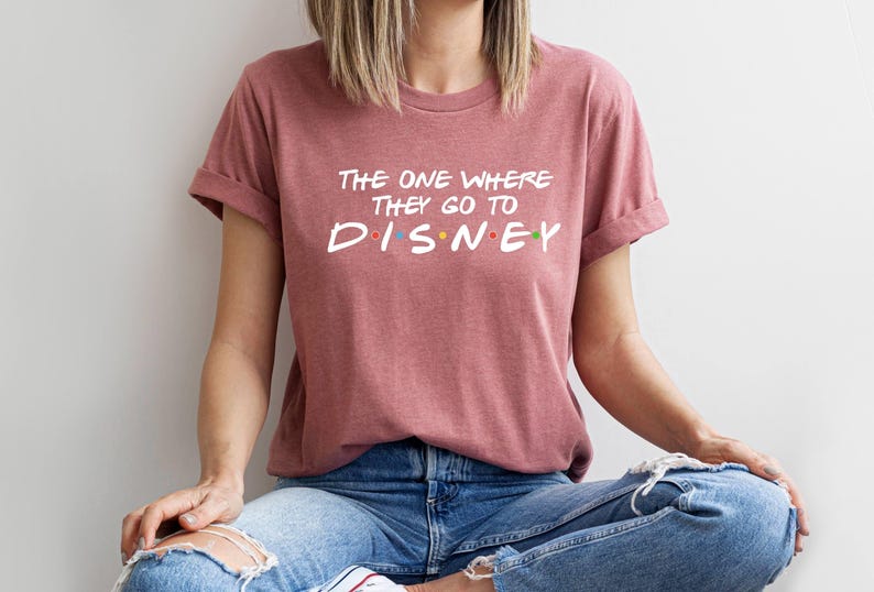 Pu&ograve; includere: T-shirt rosa polveroso con maniche arrotolate. Il testo bianco recita "The One Where They Go To Disney" con la parola "Disney" in lettere colorate. La persona indossa jeans blu.