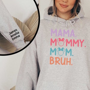K&ouml;nnte beinhalten: Grauer Kapuzenpullover mit dem Text "MAMA. MOMMY. MOM. BRUH." in bunten Buchstaben mit einem pinkfarbenen, blauen und gr&uuml;nen Mickey-Mouse-Kopf-Motiv. Der &Auml;rmel ist mit den Namen "James, Amelia, Emma" in Schwarz bestickt.