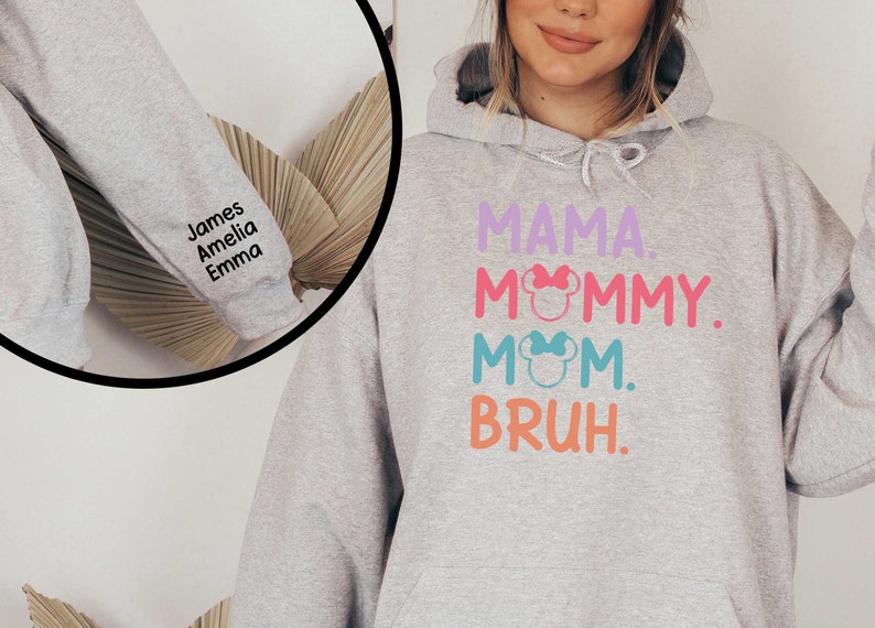 K&ouml;nnte beinhalten: Grauer Kapuzenpullover mit dem Text "MAMA. MOMMY. MOM. BRUH." in bunten Buchstaben mit einem pinkfarbenen, blauen und gr&uuml;nen Mickey-Mouse-Kopf-Motiv. Der &Auml;rmel ist mit den Namen "James, Amelia, Emma" in Schwarz bestickt.