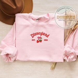 May include: Pink sweatshirt with a red embroidered design that says "Disneyland Est. 1955" with two cherries and bows.