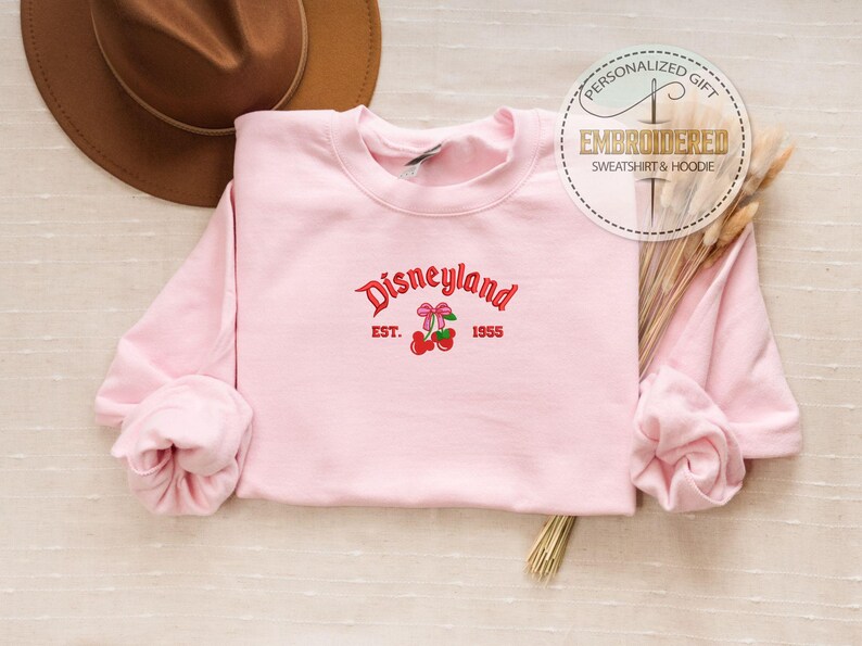 May include: Pink sweatshirt with a red embroidered design that says "Disneyland Est. 1955" with two cherries and bows.