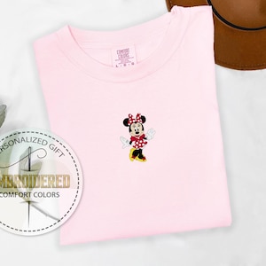 May include: Pink t-shirt with an embroidered image of Minnie Mouse in a red polka dot dress. The text "Personalized Gift" and "Embroidered Comfort Colors" is on a white circle.