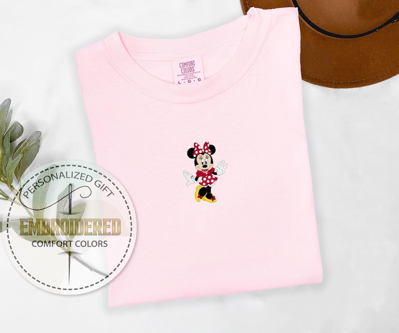 May include: Pink t-shirt with an embroidered image of Minnie Mouse in a red polka dot dress. The text "Personalized Gift" and "Embroidered Comfort Colors" is on a white circle.