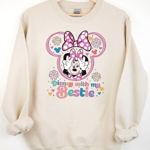 May include: A beige sweatshirt with a graphic of Minnie Mouse wearing a pink bow and holding peace signs. The graphic also includes fireworks and the text "Disney with my bestie".