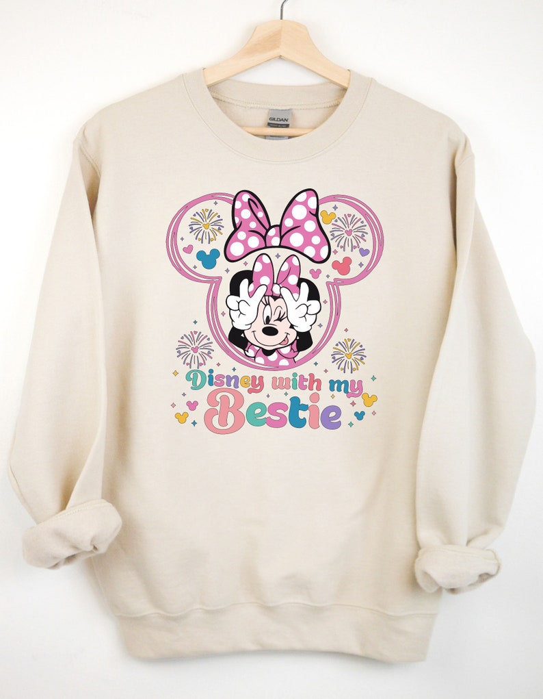 May include: A beige sweatshirt with a graphic of Minnie Mouse wearing a pink bow and holding peace signs. The graphic also includes fireworks and the text "Disney with my bestie".