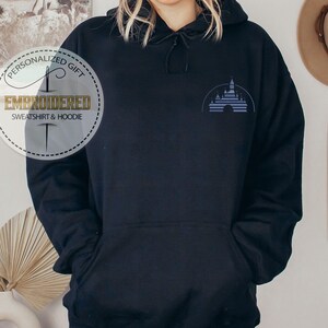 Puede incluir: Sudadera con capucha negra con una imagen bordada en azul de un castillo en la parte delantera. El texto "Regalo personalizado" y "Sudadera y sudadera con capucha bordada" est&aacute; en un c&iacute;rculo blanco con un dise&ntilde;o de aguja e hilo.