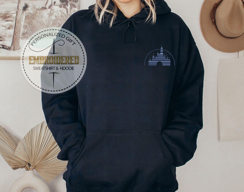 Puede incluir: Sudadera con capucha negra con una imagen bordada en azul de un castillo en la parte delantera. El texto "Regalo personalizado" y "Sudadera y sudadera con capucha bordada" est&aacute; en un c&iacute;rculo blanco con un dise&ntilde;o de aguja e hilo.