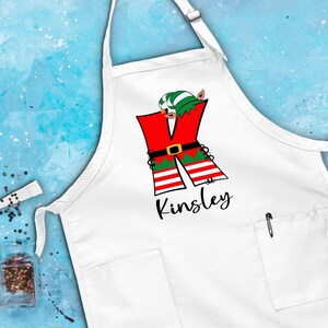 Puede incluir: Delantal de cocina blanco con una letra K de elfo roja, verde y blanca y el nombre Kinsley impreso debajo.