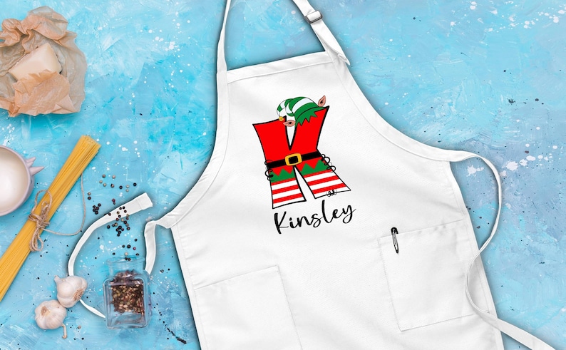 Puede incluir: Delantal de cocina blanco con una letra K de elfo roja, verde y blanca y el nombre Kinsley impreso debajo.