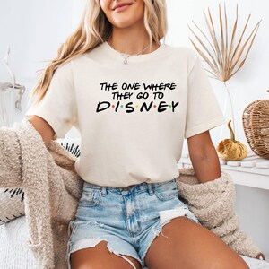 Pu&ograve; includere: T-shirt color crema con la scritta nera "THE ONE WHERE THEY GO TO DISNEY", con la parola "DISNEY" in lettere colorate. La maglietta &egrave; indossata con pantaloncini di jeans e un cardigan beige.