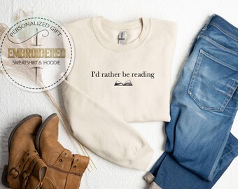 May include: Cream-colored sweatshirt with the text "I'd rather be reading" and an open book graphic. The sweatshirt is paired with blue jeans and brown boots. A circular logo reads "Embroidered Sweatshirt & Hoodie."