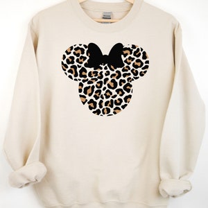 May include: A cream-colored sweatshirt with a leopard print Minnie Mouse head design. The bow is black.
