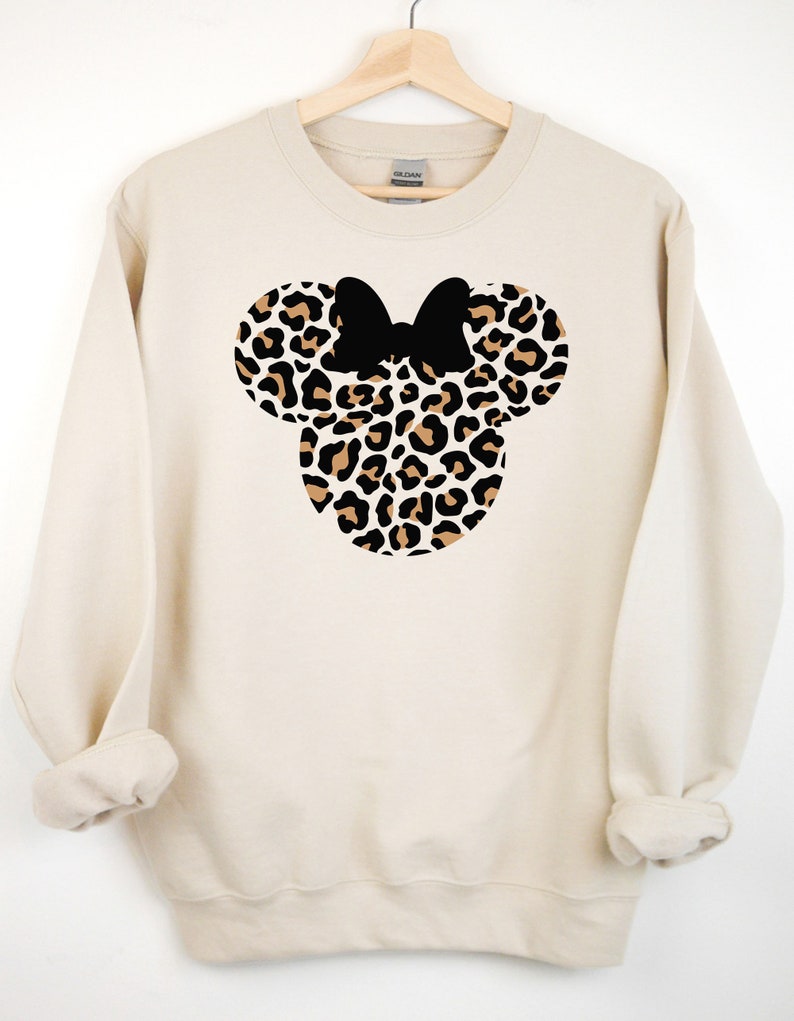 May include: A cream-colored sweatshirt with a leopard print Minnie Mouse head design. The bow is black.