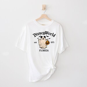 May include: White t-shirt featuring a black and white graphic of a ghost with Mickey Mouse ears holding a jack-o-lantern. The text "Disney World est 1971 FLORIDA" is printed on the shirt.