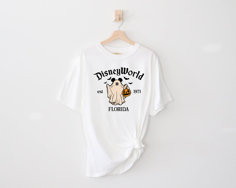May include: White t-shirt featuring a black and white graphic of a ghost with Mickey Mouse ears holding a jack-o-lantern. The text "Disney World est 1971 FLORIDA" is printed on the shirt.