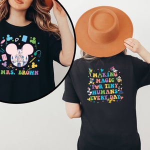 Puede incluir: Camiseta negra con un dise&ntilde;o colorido. La parte delantera presenta una cabeza de Mickey Mouse rosa, un castillo y el texto "MRS. BROWN". La parte trasera tiene el texto "MAKING MAGIC FOR TINY HUMANS EVERY DAY" con personajes de dibujos animados.
