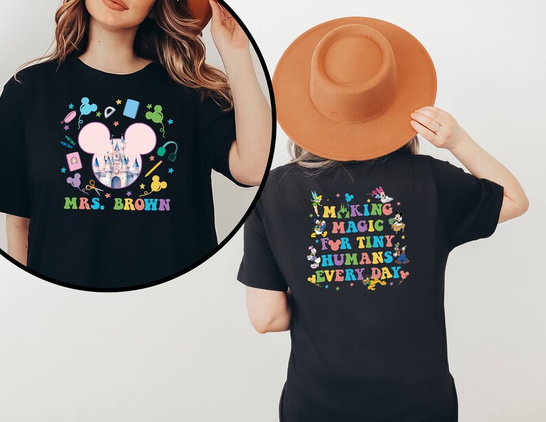 Puede incluir: Camiseta negra con un dise&ntilde;o colorido. La parte delantera presenta una cabeza de Mickey Mouse rosa, un castillo y el texto "MRS. BROWN". La parte trasera tiene el texto "MAKING MAGIC FOR TINY HUMANS EVERY DAY" con personajes de dibujos animados.