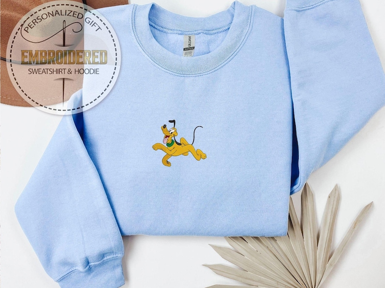 May include: Light blue sweatshirt with an embroidered Pluto graphic. The yellow dog is in a running pose with his tongue out. The text "EMBROIDERED SWEATSHIRT & HOODIE" is in a circle on the upper left.