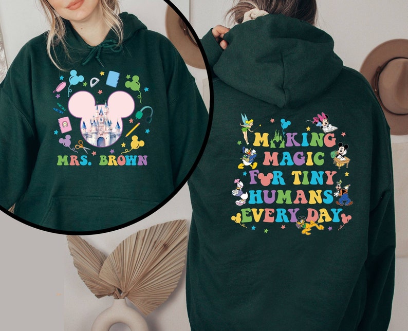 Puede incluir: Sudadera con capucha verde oscuro con un dise&ntilde;o colorido que presenta un castillo, orejas de Mickey Mouse y el texto "MRS. BROWN" en la parte delantera. La parte posterior de la sudadera dice "MAKING MAGIC FOR TINY HUMANS EVERY DAY" con personajes de dibujos animados.