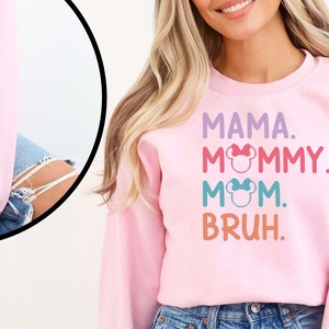 K&ouml;nnte beinhalten: Rosafarbener Pullover mit dem Text "MAMA. MOMMY. MOM. BRUH." in bunten Buchstaben mit einem Mickey-Mouse-Kopf-Motiv auf jedem Wort. Der &Auml;rmel ist mit den Namen "James, Amelia, Emma" in Schwarz bestickt.