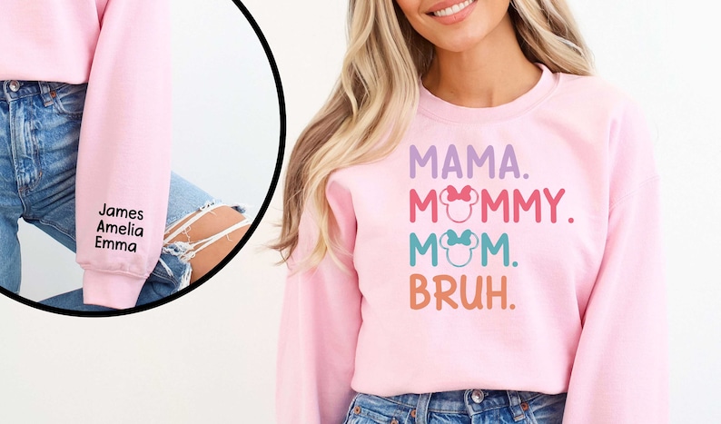 K&ouml;nnte beinhalten: Rosafarbener Pullover mit dem Text "MAMA. MOMMY. MOM. BRUH." in bunten Buchstaben mit einem Mickey-Mouse-Kopf-Motiv auf jedem Wort. Der &Auml;rmel ist mit den Namen "James, Amelia, Emma" in Schwarz bestickt.