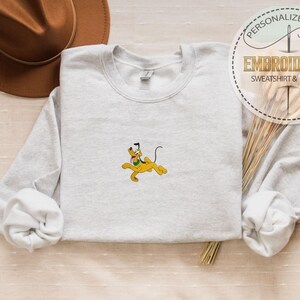 May include: A light gray sweatshirt with a yellow Pluto cartoon character embroidered on the front. The sweatshirt has long sleeves and a crew neck. The image also includes a brown hat and text that says "PERSONALIZED GIFT EMBROIDERED SWEATSHIRT & HOODIE."