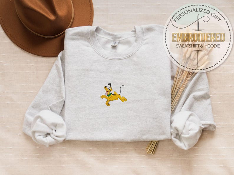 May include: A light gray sweatshirt with a yellow Pluto cartoon character embroidered on the front. The sweatshirt has long sleeves and a crew neck. The image also includes a brown hat and text that says "PERSONALIZED GIFT EMBROIDERED SWEATSHIRT & HOODIE."