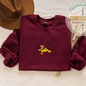 May include: A maroon sweatshirt with a small embroidered Pluto design in yellow, green, and black. The sweatshirt has long sleeves and a crew neck. The image also includes a brown hat and text that reads "PERSONALIZED GIFT EMBROIDERED SWEATSHIRT & HOODIE".