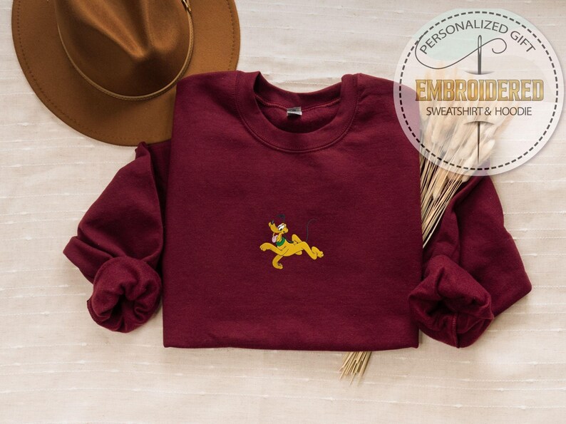 May include: A maroon sweatshirt with a small embroidered Pluto design in yellow, green, and black. The sweatshirt has long sleeves and a crew neck. The image also includes a brown hat and text that reads "PERSONALIZED GIFT EMBROIDERED SWEATSHIRT & HOODIE".