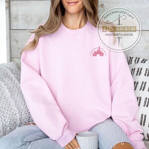 Puede incluir: Una sudadera rosa con una peque&ntilde;a imagen bordada del castillo de Disney en el pecho izquierdo. El texto "Regalo personalizado" y "Sudadera y sudadera con capucha bordadas" est&aacute; en un c&iacute;rculo dorado sobre el hombro del modelo.