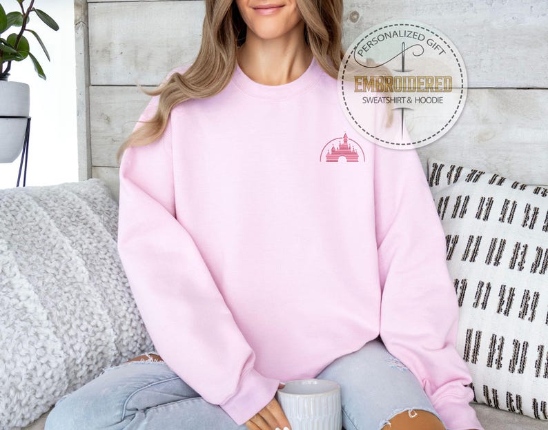 Puede incluir: Una sudadera rosa con una peque&ntilde;a imagen bordada del castillo de Disney en el pecho izquierdo. El texto "Regalo personalizado" y "Sudadera y sudadera con capucha bordadas" est&aacute; en un c&iacute;rculo dorado sobre el hombro del modelo.