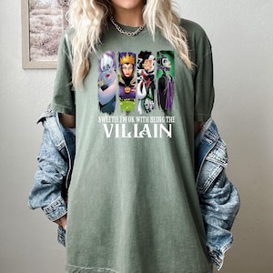 Pu&ograve; includere: T-shirt verde oliva con una grafica di quattro cattivi dei cartoni animati e la scritta "Sweetie I'm Ok With Being The Villain". La maglietta &egrave; indossata da una persona con i capelli biondi e una giacca di jeans.