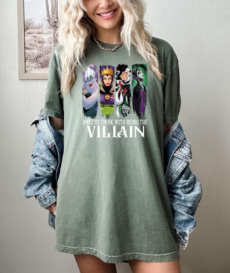 Pu&ograve; includere: T-shirt verde oliva con una grafica di quattro cattivi dei cartoni animati e la scritta "Sweetie I'm Ok With Being The Villain". La maglietta &egrave; indossata da una persona con i capelli biondi e una giacca di jeans.