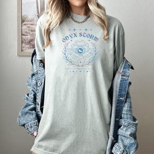 May include: A light green t-shirt with a graphic design of a flower with an eye in the center. The text "ONYX STORM" is above the flower and the text "I will be the storm, I will save" is below the flower.