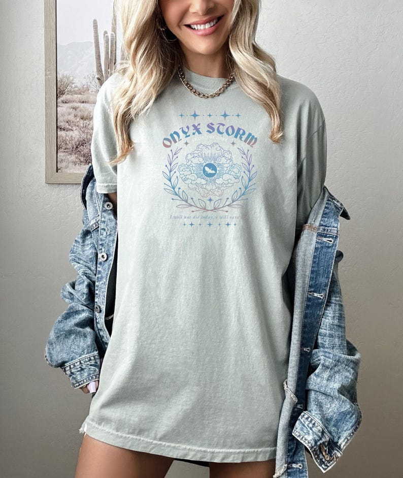 May include: A light green t-shirt with a graphic design of a flower with an eye in the center. The text "ONYX STORM" is above the flower and the text "I will be the storm, I will save" is below the flower.