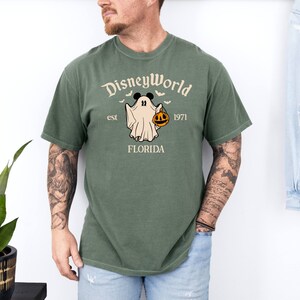 May include: A green t-shirt featuring a white graphic of a ghost holding a jack-o'-lantern. The graphic also includes the text "Disney World est 1971 Florida".