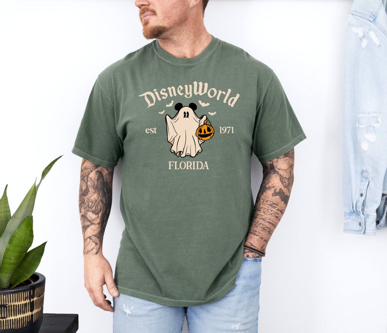May include: A green t-shirt featuring a white graphic of a ghost holding a jack-o'-lantern. The graphic also includes the text "Disney World est 1971 Florida".