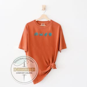 May include: An orange t-shirt with turquoise embroidered sea-themed designs, including a seashell, crab, and seaweed. The shirt is hanging on a wooden hanger. The text "PERSONALIZED GIFT EMBROIDERED COMFORT COLORS" is printed on a circular sticker.