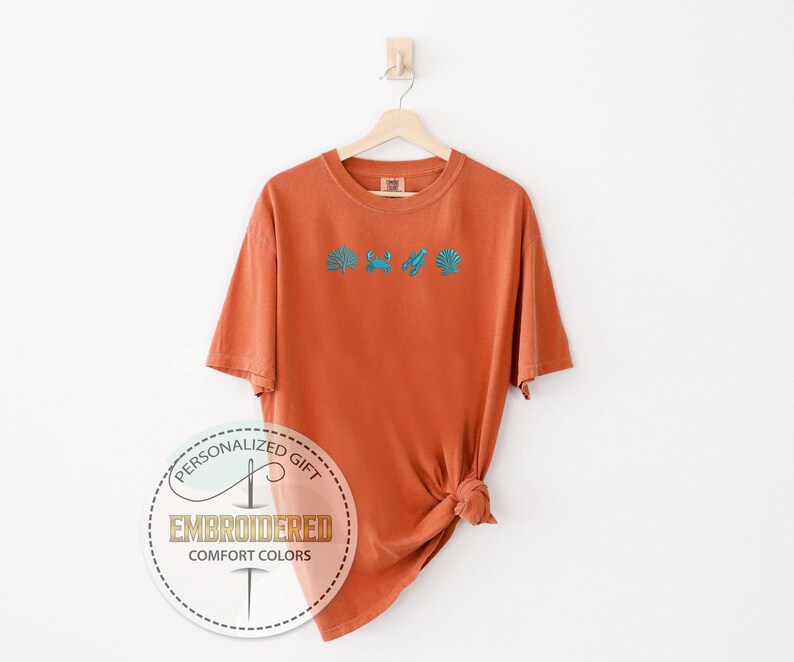 May include: An orange t-shirt with turquoise embroidered sea-themed designs, including a seashell, crab, and seaweed. The shirt is hanging on a wooden hanger. The text "PERSONALIZED GIFT EMBROIDERED COMFORT COLORS" is printed on a circular sticker.