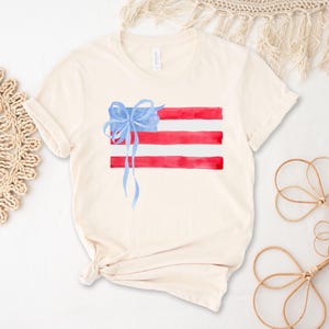 May include: A white t-shirt featuring a watercolour design of an American flag with a blue bow on top.