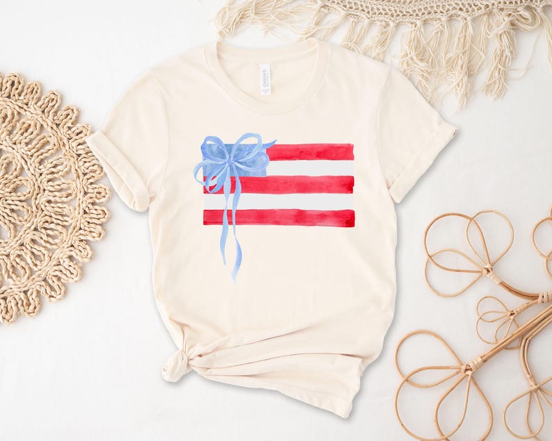 May include: A white t-shirt featuring a watercolour design of an American flag with a blue bow on top.