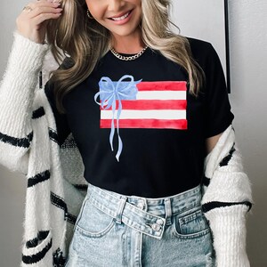 May include: A black t-shirt featuring a watercolour design of an American flag with a blue bow on top. The shirt is being worn by a person with long blonde hair and a white cardigan.