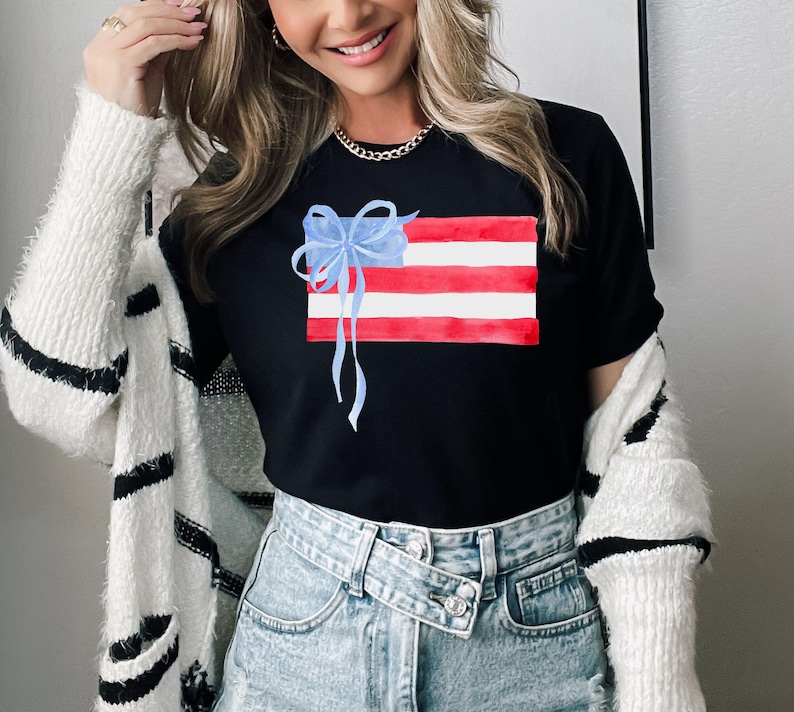 May include: A black t-shirt featuring a watercolour design of an American flag with a blue bow on top. The shirt is being worn by a person with long blonde hair and a white cardigan.