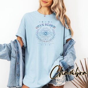 May include: A light blue t-shirt with a graphic design of a flower with a blue eye in the center. The text "Onyx Storm" is above the flower and the text "I will not live in fear, I will own him" is below the flower.