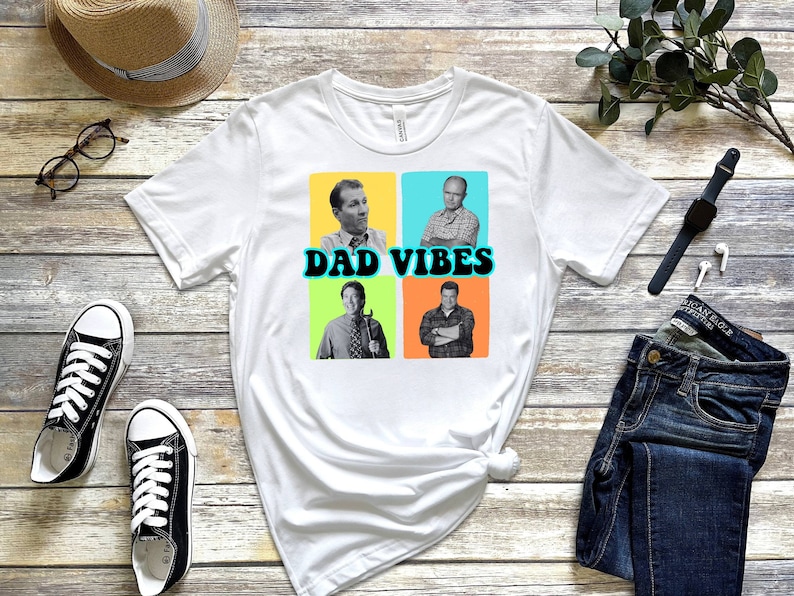 May include: White t-shirt featuring a graphic of four men in colourful squares with the text "DAD VIBES" in a retro font.