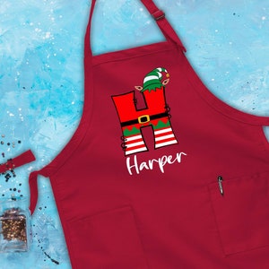 Puede incluir: Delantal rojo de Navidad con un bolsillo blanco y un dise&ntilde;o personalizado con la letra "H" vestida de elfo con el nombre "Harper" debajo.
