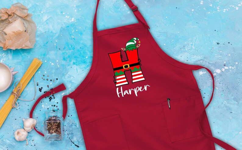 Puede incluir: Delantal rojo de Navidad con un bolsillo blanco y un dise&ntilde;o personalizado con la letra "H" vestida de elfo con el nombre "Harper" debajo.