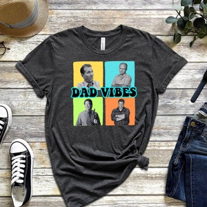 May include: A dark grey t-shirt featuring a graphic of four men in a collage. The men are wearing casual clothing and have a retro look. The text "DAD VIBES" is printed in a blue and yellow font.