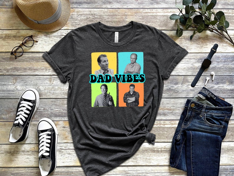 May include: A dark grey t-shirt featuring a graphic of four men in a collage. The men are wearing casual clothing and have a retro look. The text "DAD VIBES" is printed in a blue and yellow font.