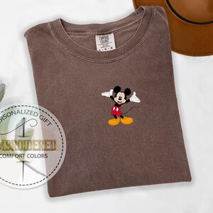 May include: A brown t-shirt with a small embroidered image of Mickey Mouse in the center. The shirt is a unisex style and has a crew neck.